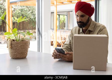 Uomo indiano medio adulto in turbante seduto a Kitchen Island che controlla smartphone vicino alla pianta, scatola sigillata Foto Stock