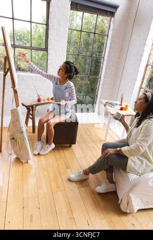 Madre asiatica e figlia adolescente che tiene in mano una tazza di ceramica e dipinge su un cavalletto in legno in studio d'arte Foto Stock