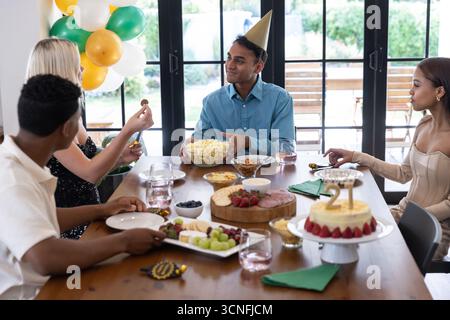 Diversi amici che festeggiano il 21° compleanno intorno al tavolo da pranzo a casa con torta e popcorn Foto Stock