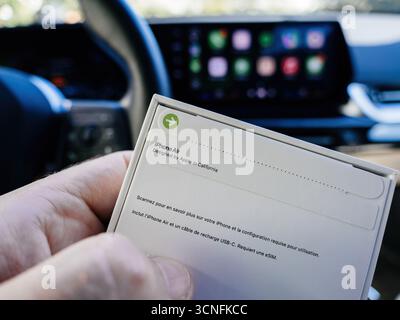 Parigi, Francia - 19 settembre 2025: Tenere a mano il retro della confezione dell'iPhone Air con logo e dettagli QR, con la schermata del cruscotto CarPlay nella Foto Stock