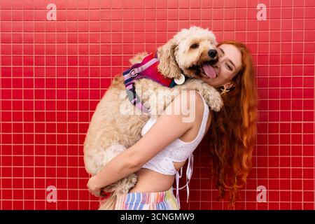 Redhead Woman abbraccia il suo gioioso cane da doodle, condividendo un caldo momento di affetto e compagnia contro una vivace parete piastrellata, giocosa estate all'aperto Foto Stock