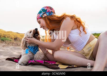 Giovane donna dai capelli rossi con una bandana colorata che accarezza il suo cane doodle felice che indossa una bandana blu mentre si rilassa su una coperta nella sabbia durante un su Foto Stock
