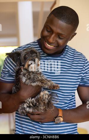 Uomo afro-americano che tiene un piccolo cane grigio e nero in una casa molto illuminata Foto Stock