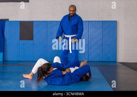 Istruttore afro-americano che guida le studentesse adolescenti che affrontano gi su tappeti nel dojo Foto Stock