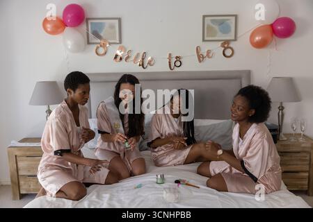 Diverse amiche che indossano abiti sul letto in camera da letto per festeggiare il addio al nubilato con palloncini Foto Stock