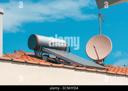 Scaldabagno solare sul tetto e parabola satellitare installati su piastrelle di terracotta sotto il cielo azzurro. Sistema di energia rinnovabile ecocompatibile su uso residenziale Foto Stock