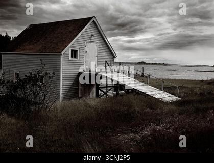 Un rustico capannone di barche si trova vicino a un tranquillo canale della nuova Scozia, circondato da erba alta e un cielo nuvoloso. La struttura è dotata di una semplice rampa che conduce a. Foto Stock