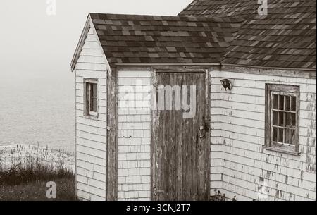 Un edificio rustico sorge accanto all'acqua, circondato da erba alta. La struttura presenta un esterno bianco sbiadito e una porta in legno usurato, protetta in una S. Foto Stock