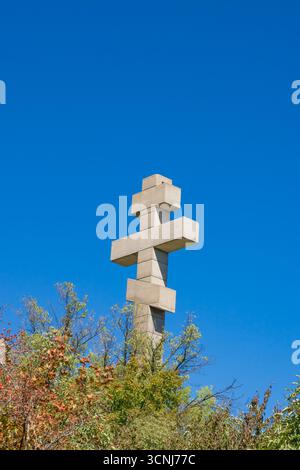 Il grande monumento a croce ortodossa si erge alto sopra colorati alberi autunnali, sagomato contro un cielo blu limpido, a simboleggiare la fede e la spiritualità in un Foto Stock