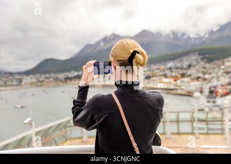 Una donna bionda cattura il suggestivo porto e lo skyline montuoso di Ushuaia con il suo smartphone dal ponte di una nave da crociera. Foto Stock