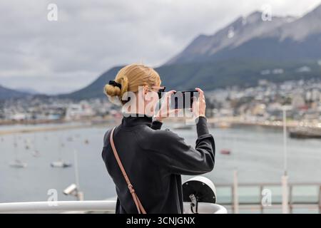 Una donna bionda cattura il suggestivo porto e lo skyline montuoso di Ushuaia con il suo smartphone dal ponte di una nave da crociera. Foto Stock