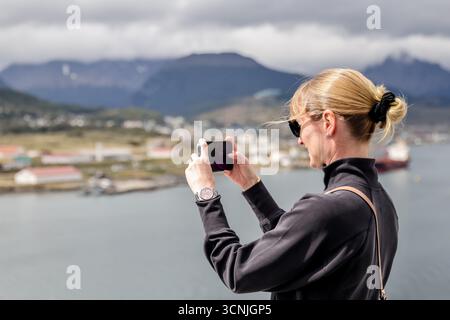 Una donna bionda cattura il suggestivo porto e lo skyline montuoso di Ushuaia con il suo smartphone dal ponte di una nave da crociera. Foto Stock