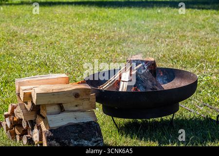 Caminetto all'aperto con fiamme e tronchi in fiamme, circondato da legna da ardere ben accatastata su lussureggiante erba verde, che offre un'atmosfera calda e invitante Foto Stock