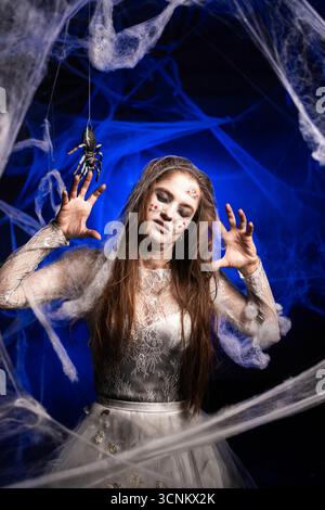 Donna inquietante con orribile trucco zombie di Halloween in abito bianco su sfondo blu circondato da ragnatela. Persona ossessionata dal diavolo. Horro Foto Stock