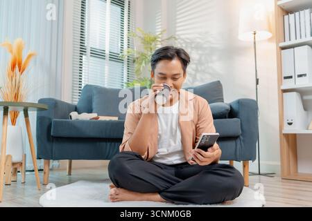 Un giovane uomo siede comodamente sul pavimento con le gambe incrociate, sorridendo mentre utilizza uno smartphone in un salotto accogliente. L'atmosfera rilassata si distingue Foto Stock
