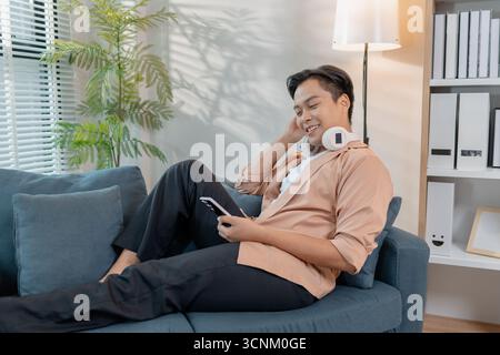 Un giovane uomo si rilassa a casa, sorridendo mentre usa il suo smartphone. Ama trascorrere il tempo libero con le cuffie intorno al collo, seduto comodamente e comodo Foto Stock