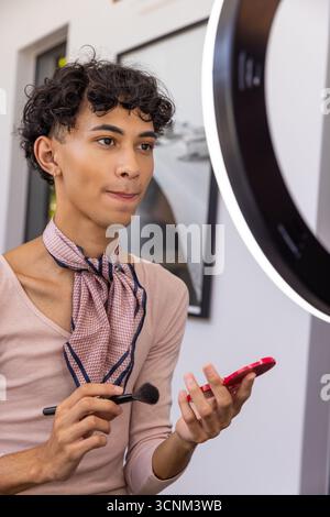 Uomo che tiene in mano uno specchio compatto e applica il trucco in studio con luce anulare vicino alla fotografia incorniciata Foto Stock