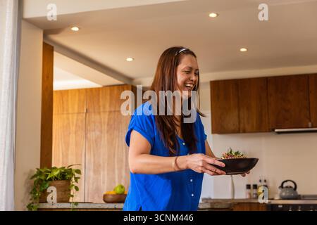 Donna asiatica matura che indossa un vestito blu in piedi in cucina tenendo insalata ciotola accanto alla pianta in vaso Foto Stock