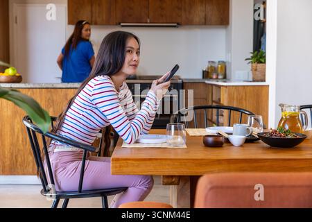 Madre asiatica che prepara il succo al bancone mentre la figlia guarda lo smartphone al tavolo da pranzo Foto Stock