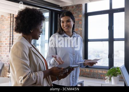Diverse colleghe discutono di strategia, tenendo tablet e documenti in ufficio con un notebook Foto Stock