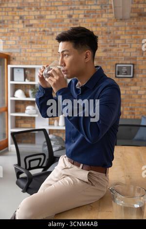 Uomo asiatico che indossa una camicia seduto alla scrivania in ufficio bevendo acqua dal vetro accanto alla caraffa Foto Stock