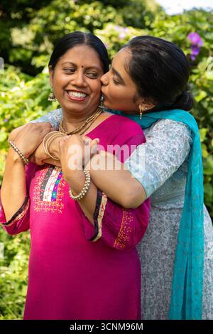 Madre e figlia indiane abbracciano e baciano le guance in giardino con kurta ricamati Foto Stock