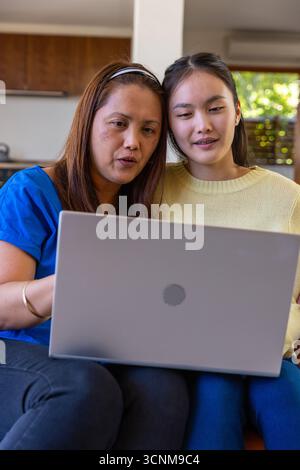 Madre e figlia asiatica sedute sul divano di casa vicino alla cucina imparando insieme sul laptop Foto Stock