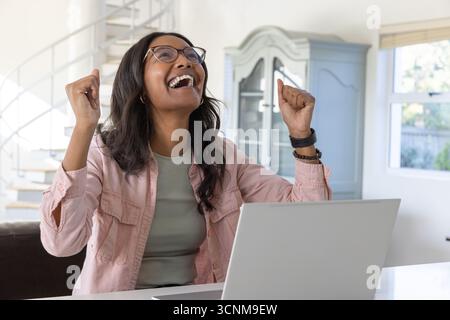 Donna afro-americana che celebra il successo seduto in casa con un laptop e vista sul giardino Foto Stock