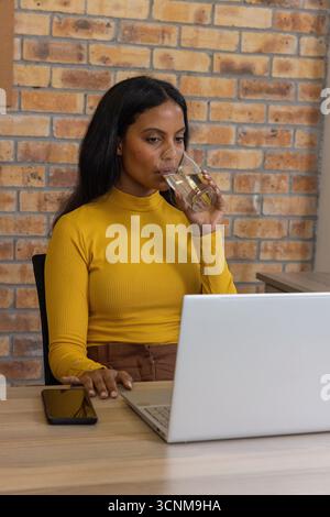 Donna seduta a una scrivania di legno in un ufficio domestico con un laptop color argento e sorseggiando acqua Foto Stock