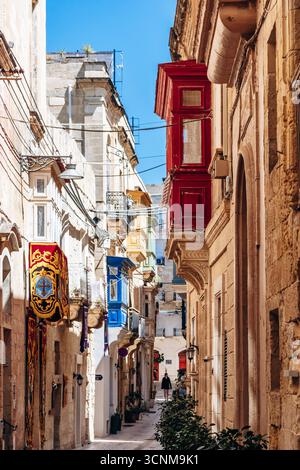 BIRGU, MALTA - 11 APRILE 2025: Tradizionale via maltese con balconi in legno colorati, bandiere maltesi rosse e bianche e storico edificio in pietra calcarea Foto Stock