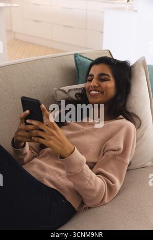 Donna asiatica sdraiata sul divano beige con una felpa rosa nella lounge moderna utilizzando lo smartphone Foto Stock