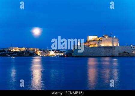 Fort St. Angelo illuminato sorge sul Grand Harbour al crepuscolo di Malta, i suoi bastioni calcarei che brillano sotto il cielo blu della sera e la luna piena, Foto Stock