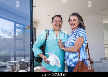 Coppia senior in piedi all'ingresso con zaino nero che regge cappello da sole e borsa in pelle Foto Stock