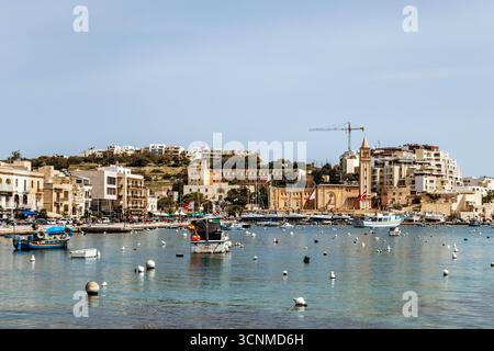 Vista panoramica sul lungomare di Marsaskala con edifici colorati, barche e acque turchesi limpide in un giorno di primavera soleggiato, Malta, Europa Foto Stock