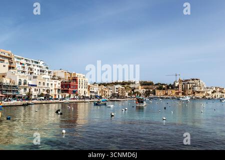 Vista panoramica sul lungomare di Marsaskala con edifici colorati, barche e acque turchesi limpide in un giorno di primavera soleggiato, Malta, Europa Foto Stock