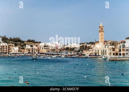Vista panoramica sul lungomare di Marsaskala con edifici colorati, barche e acque turchesi limpide in un giorno di primavera soleggiato, Malta, Europa Foto Stock