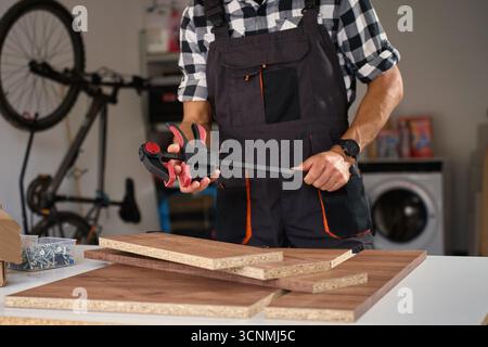 Primo piano di artigiani in tute con morsetto per preparare pannelli di legno per l'assemblaggio di mobili. Falegname che assembla pannelli di mobili in legno in officina Foto Stock