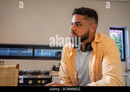Un uomo adulto seduto in cucina con le cuffie che guardano il piano cottura e le finestre Foto Stock