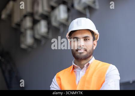 Giovane uomo barbuto in piedi all'aperto, indossando un elmetto bianco e un giubbotto di sicurezza arancione, che rappresenta la costruzione, l'ingegneria, la sicurezza e una professoressa Foto Stock