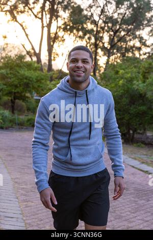 Uomo afroamericano che cammina lungo il sentiero lastricato nel parco al crepuscolo indossando felpe con cappuccio e pantaloncini neri Foto Stock