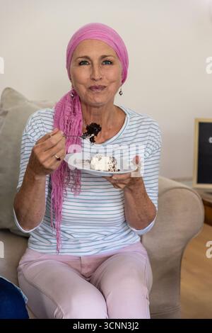 Donna anziana che indossa il velo seduto sul divano a casa tenendo la forchetta sopra la torta sul piatto Foto Stock