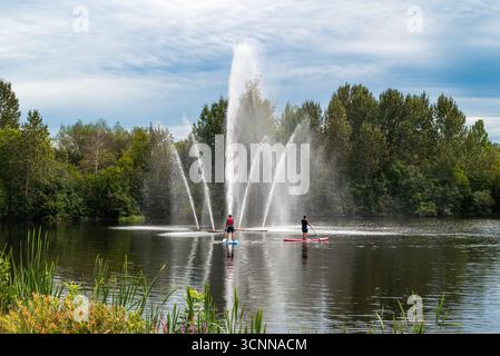 Sali a paddleboard di fronte a una grande fontana d'acqua su uno stagno circondato da alberi a Saint-Georges-de-Beauce, Quebec, Canada Foto Stock