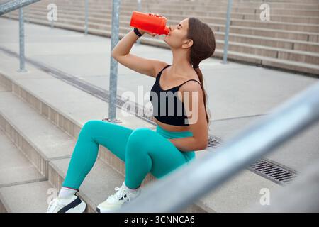Una giovane donna in abbigliamento sportivo si prende una pausa su una serie di scale all'aperto, sorseggiando da una bottiglia d'acqua dopo una sessione di allenamento energica in mezzo a un vivace Foto Stock