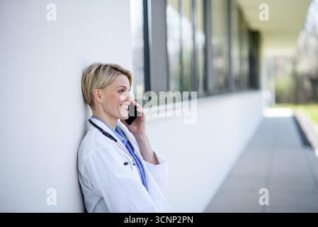 Giovane medico donna che fa una pausa, seduta sulla panchina di fronte all'ospedale e fa la chiamata. Foto Stock