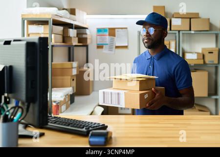 Giovane adulto di colore che indossa un cappello che regge una pila di pacchi, in piedi al bancone del magazzino che lavora con pacchi vicino al monitor del computer circondato da scatole di cartone e materiali di spedizione Foto Stock