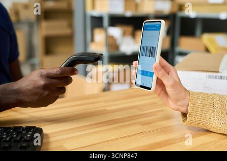 Giovane donna caucasica adulta che tiene in mano uno smartphone che visualizza codici a barre mentre l'uomo nero utilizza un lettore di codici a barre portatile, che interagisce presso una scrivania di legno in magazzino o in una struttura di spedizione Foto Stock