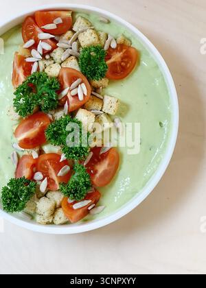 zuppa verde cremosa presentata in una ciotola bianca pulita, splendidamente decorata con un mix colorato e strutturato di metà di pomodoro ciliegino rosso brillante, croccante Foto Stock