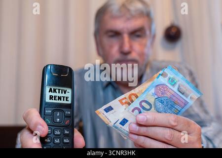 Germania - 21 settembre 2025: FOTOMONTAGGIO, un uomo anziano ha contanti e un telefono con l'annuncio: Aktivrente. La foto simboleggia il reddito aggiuntivo e la sicurezza finanziaria in pensione *** FOTOMONTAGE, Ein älterer Mann hält Bargeld und ein Telefon mit der Anzeige: Aktivrente. DAS foto steht symbolisch für zusätzliche Einkünfte und finanzielle Absicherung im Ruhestand Foto Stock