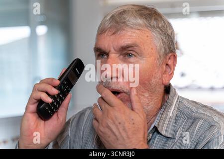 Germania - 21 settembre 2025: Un uomo anziano tiene un telefono all'orecchio sotto shock. L'immagine illustra il pericolo di chiamate d'emergenza e truffe telefoniche, che sono progettate per turbare e spaventare gli anziani. *** Ein älterer Mann hält schockiert ein Telefon am Ohr. DAS Bild verdeutlicht die Gefahr von Schockanrufen und Telefonbetrug, die gezielt Senioren verunsichern und in Angst versetzen sollen. Foto Stock