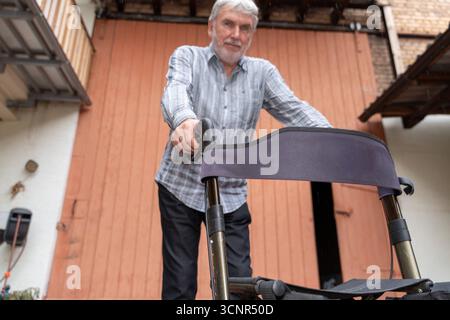 Germania - 21 settembre 2025: Un uomo anziano si trova di fronte a un edificio, aggrappandosi al suo camminatore. *** Ein älterer Mann steht vor einem Gebäude und hält sich un festival di Rollator con la Senna. Foto Stock